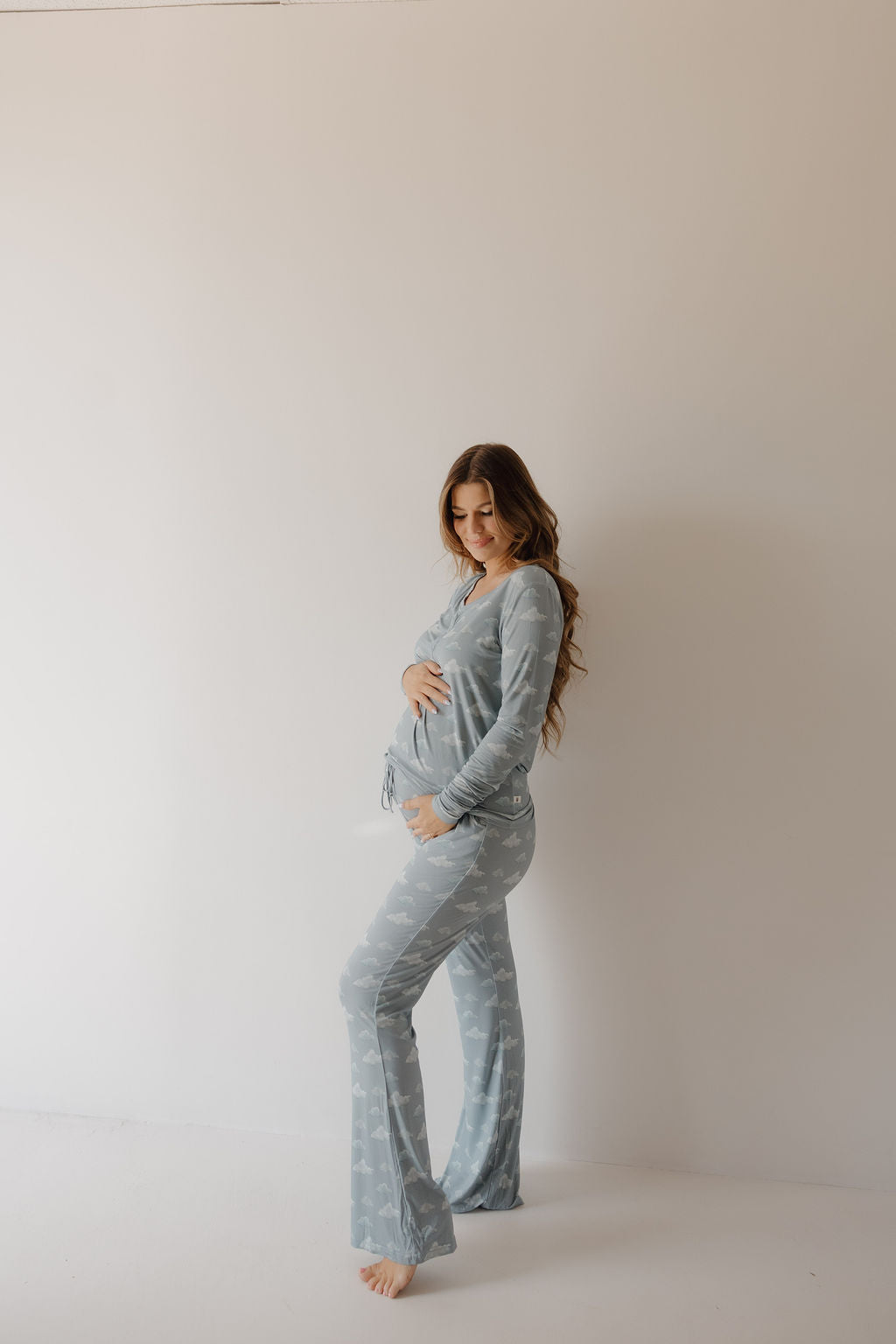 A pregnant woman with long brown hair stands barefoot against a light background, smiling down at her baby bump while wearing Kendy 3’s Women's Bamboo Pajama | Duke, made from breathable bamboo fabric.