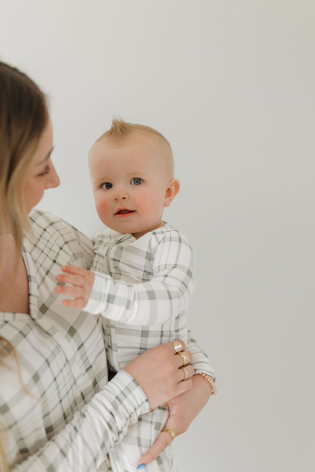 A woman lovingly holds a baby with a small mohawk, both dressed in matching "Bamboo Zip Pajamas | Picnic Plaid" by forever french baby, against a plain, light-colored background.