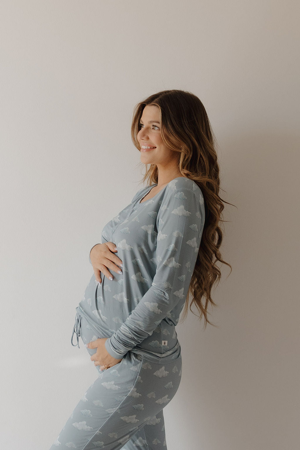 A smiling pregnant woman with long, wavy hair gently cradles her belly while wearing Kendy 3 Women's Bamboo Pajama | Duke in a light blue cloud print, enjoying the breathable comfort against a plain background.