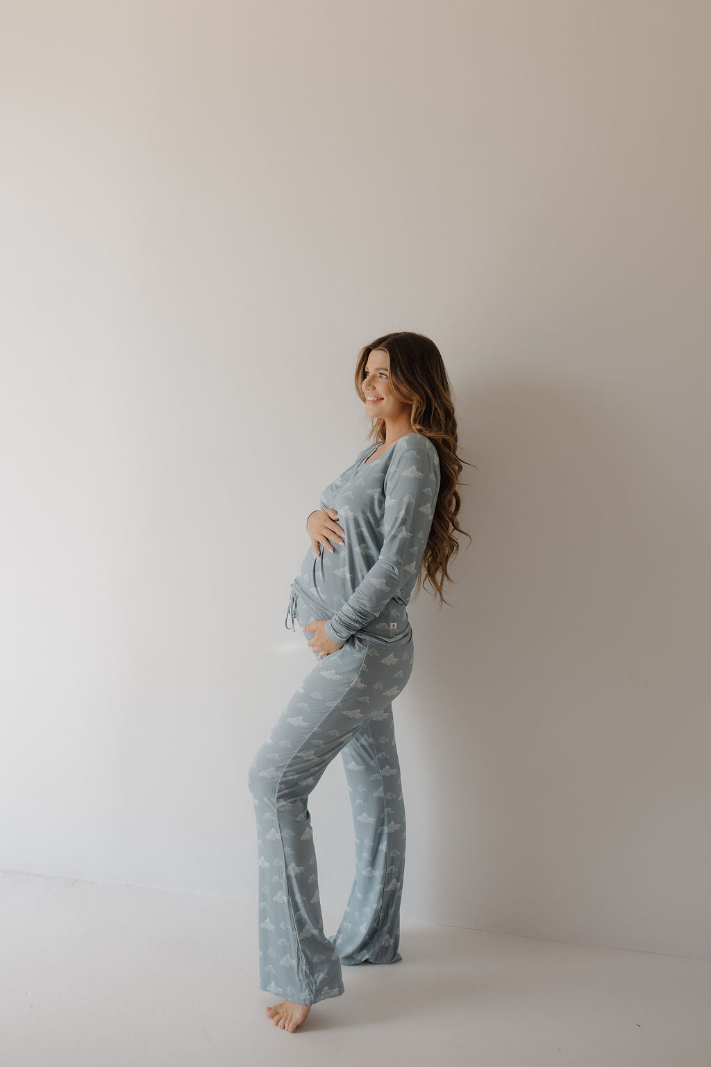 A woman in Kendy 3’s Women's Bamboo Pajama | Duke, featuring a light blue hue with a subtle white pattern, stands barefoot and smiles as she gently cradles her pregnant belly, enjoying the pajamas’ soft and breathable bamboo fabric.