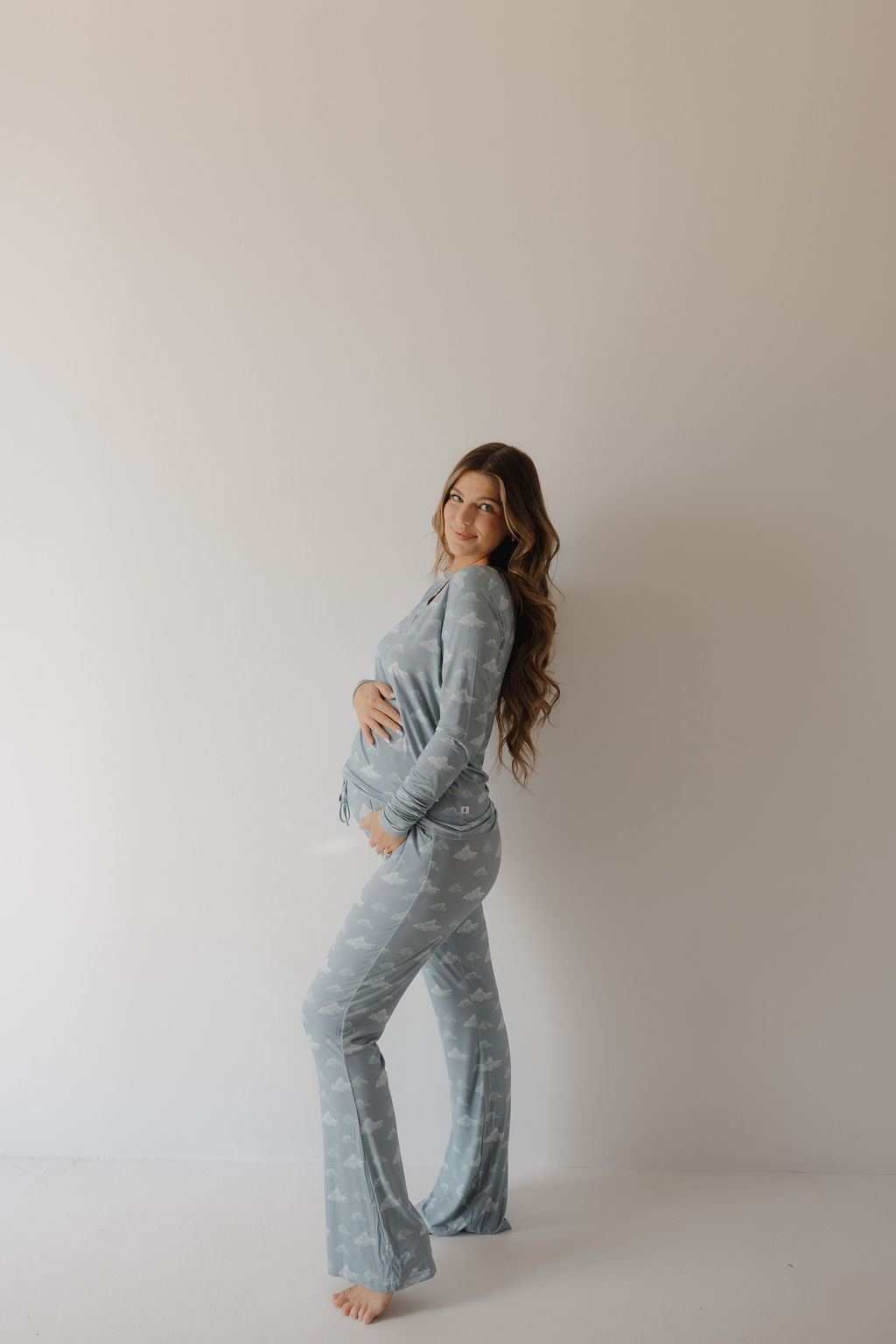 A pregnant woman with long brown hair, smiling and cradling her belly, wears Kendy 3's Women's Bamboo Pajama | Duke in light blue. Made from breathable fabric, she poses sideways against a plain, light-colored wall.