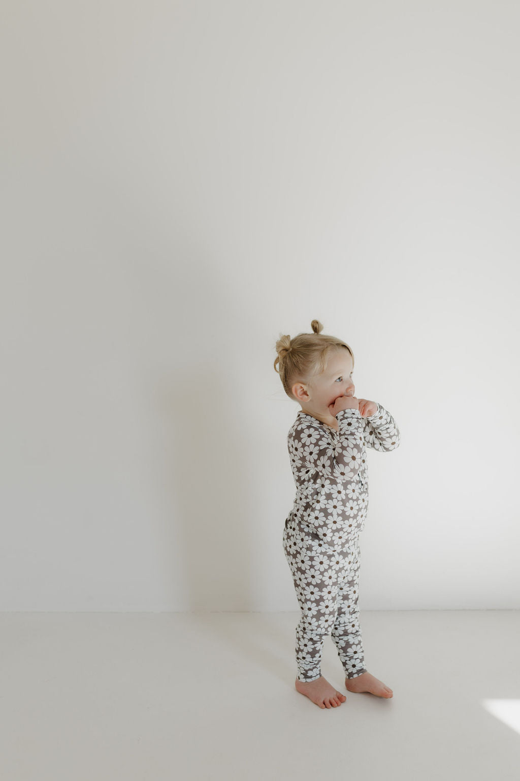 A toddler with light hair in small pigtails, wearing forever french baby's Bamboo Two Piece Pajamas | Darlin' featuring a star pattern, stands against a plain white wall. The child holds one hand near their mouth, gazing off to the side.