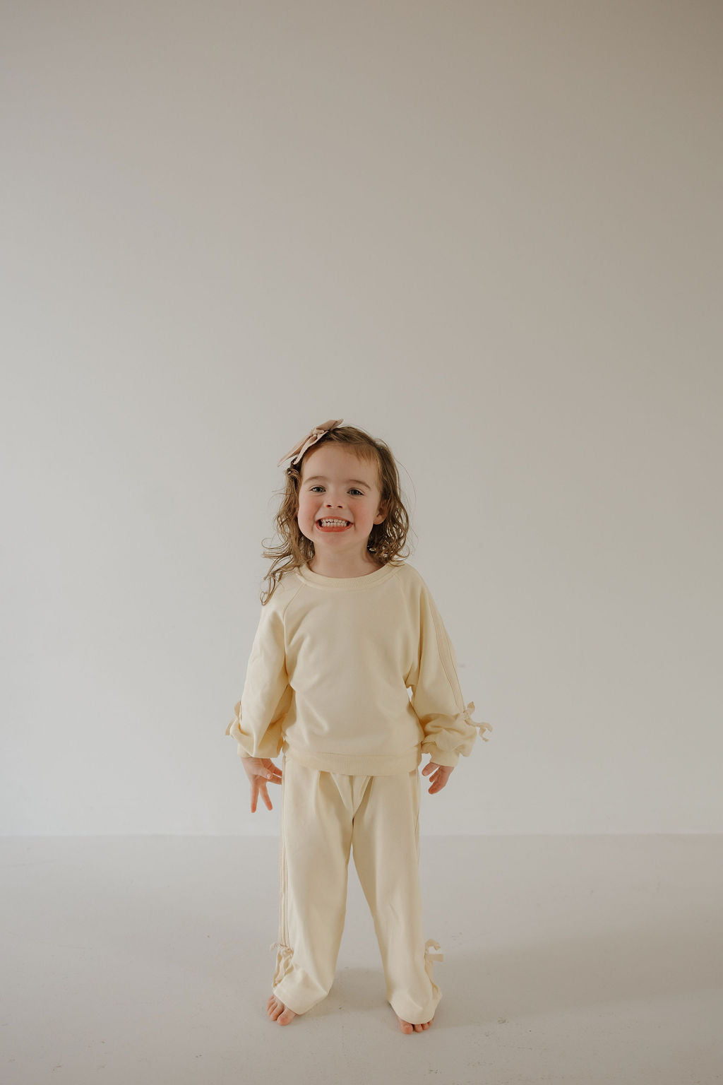 A young child stands barefoot and smiling in forever french baby’s Child's Sweat Set | Buttercream Bows, featuring a cream hue and bow details. The cozy long-sleeve top and pants stand out against the simple, light background.