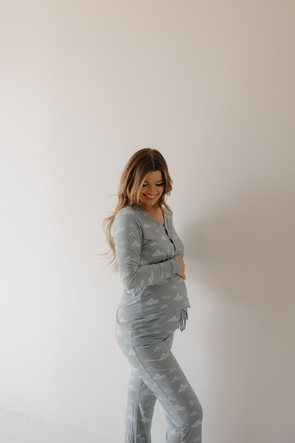 A pregnant woman stands sideways, smiling and touching her belly. She wears Kendy 3's Women's Bamboo Pajama | Duke in light blue with a white cloud pattern, featuring breathable fabric against a plain background.