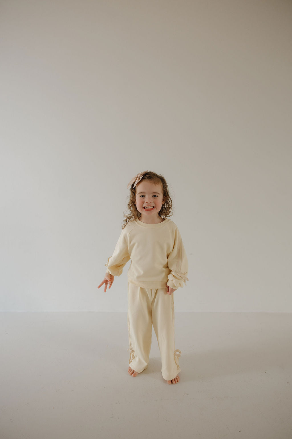 A young child with long, curly hair wears the forever french baby Child's Sweat Set in Buttercream Bows, standing barefoot against a plain light background and smiling at the camera.