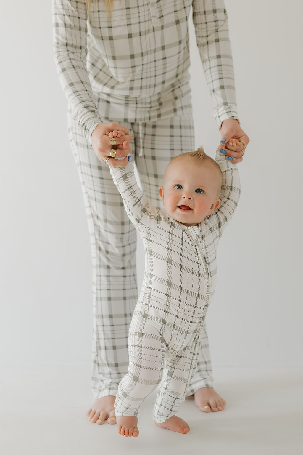 A baby in forever french baby's Bamboo Zip Pajamas | Picnic Plaid is supported by an adult, whose face isn't visible. The little one beams a joyful smile with a tiny mohawk haircut against a plain white background, showcasing the pajamas' breathable comfort.