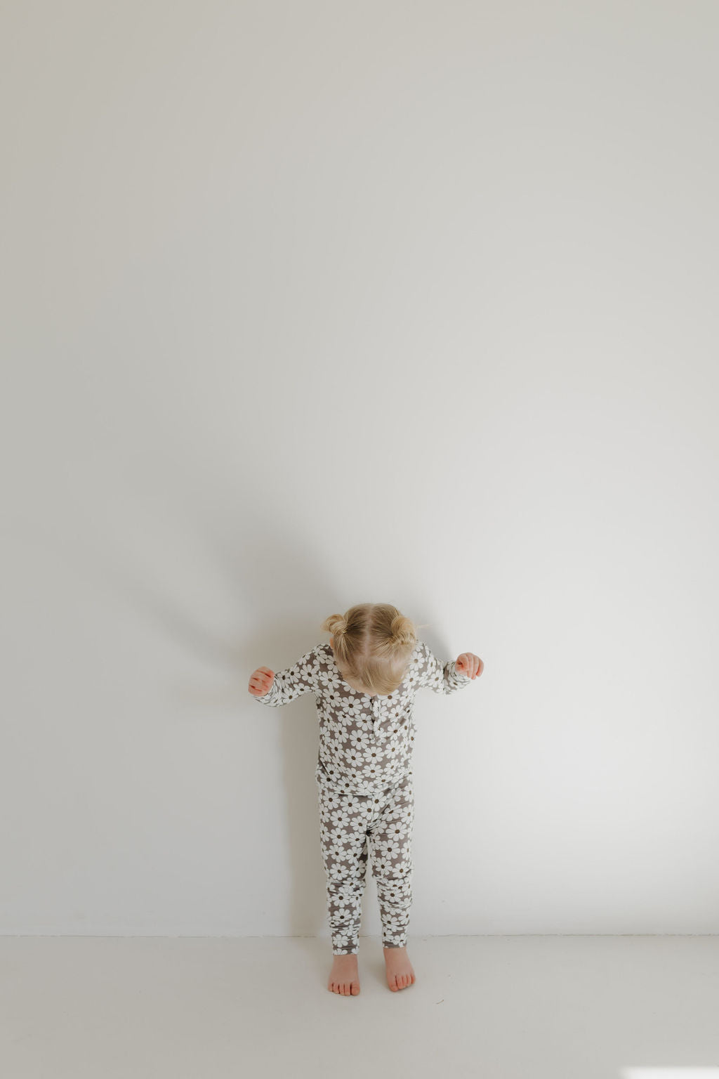 A child wearing forever french baby's Bamboo Two Piece Pajamas | Darlin' stands against a plain white wall, looking down with arms slightly outstretched. The minimalistic scene features soft lighting and a calm atmosphere.