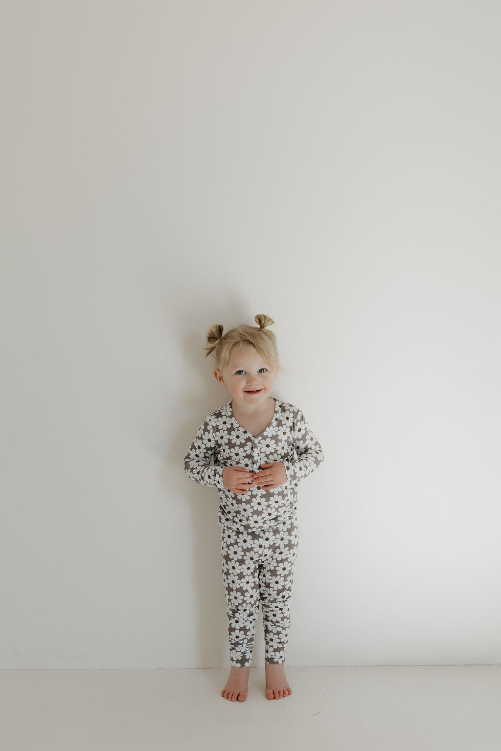 A young child with blonde hair in two buns stands barefoot against a plain white wall, wearing forever french baby's hypo-allergenic Bamboo Two Piece Pajamas | Darlin' with a black and white star pattern, holding a small object.