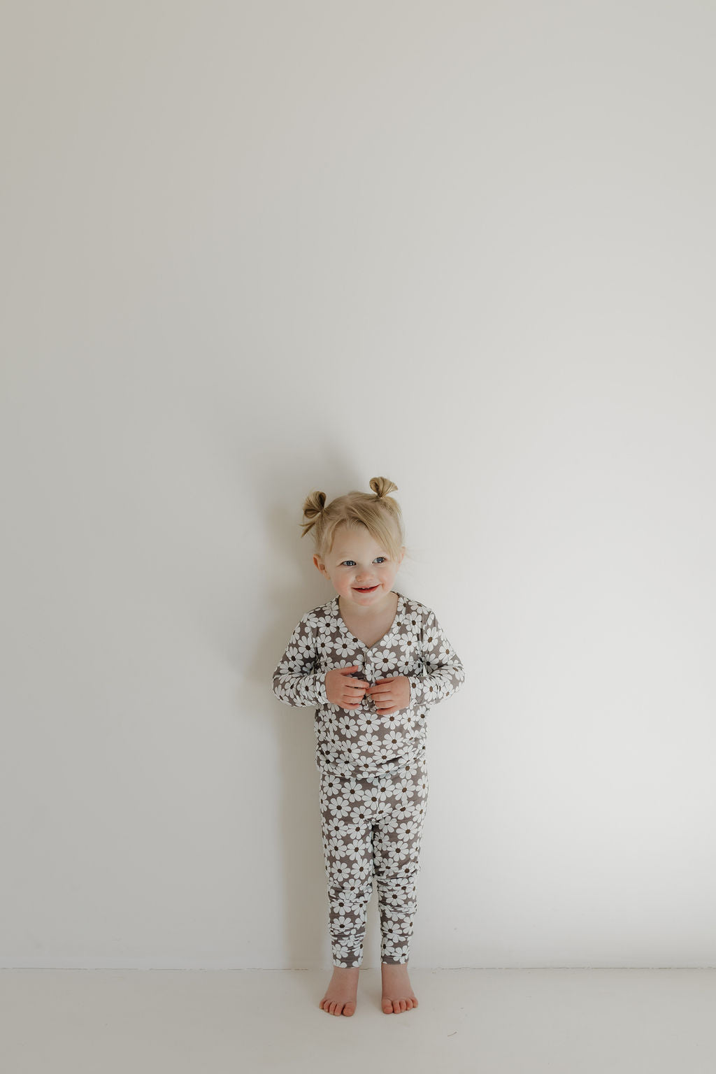 A young child with blonde hair in pigtails stands barefoot against a white wall, smiling while wearing forever french baby's hypo-allergenic Bamboo Two Piece Pajamas | Darlin'. The breathable, star-patterned long-sleeve set ensures comfort as they hold their hands together in front.