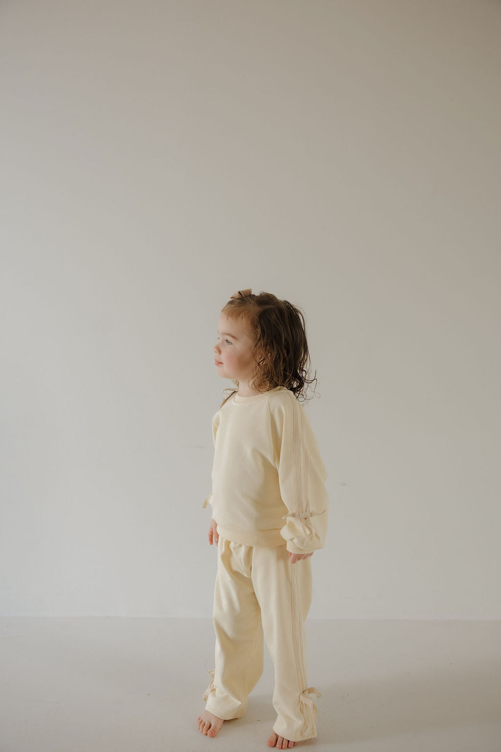 A young child with brown hair stands indoors against a light wall, wearing the Child's Sweat Set | Buttercream Bows by forever french baby—an ideal cozy choice for families seeking matching outfits.