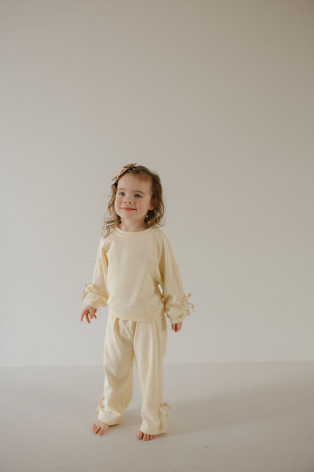 A young child with wavy hair stands barefoot against a light background, wearing forever french baby's Child's Sweat Set | Buttercream Bows with ruffle sleeve and cuff details—perfect for cozy days or matching family outfits.
