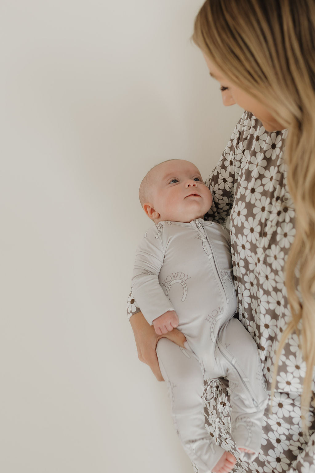 A woman in a floral-patterned outfit lovingly gazes at her baby, who is wearing light-colored, hypoallergenic Bamboo Zip Pajamas by forever french baby, against a plain background.