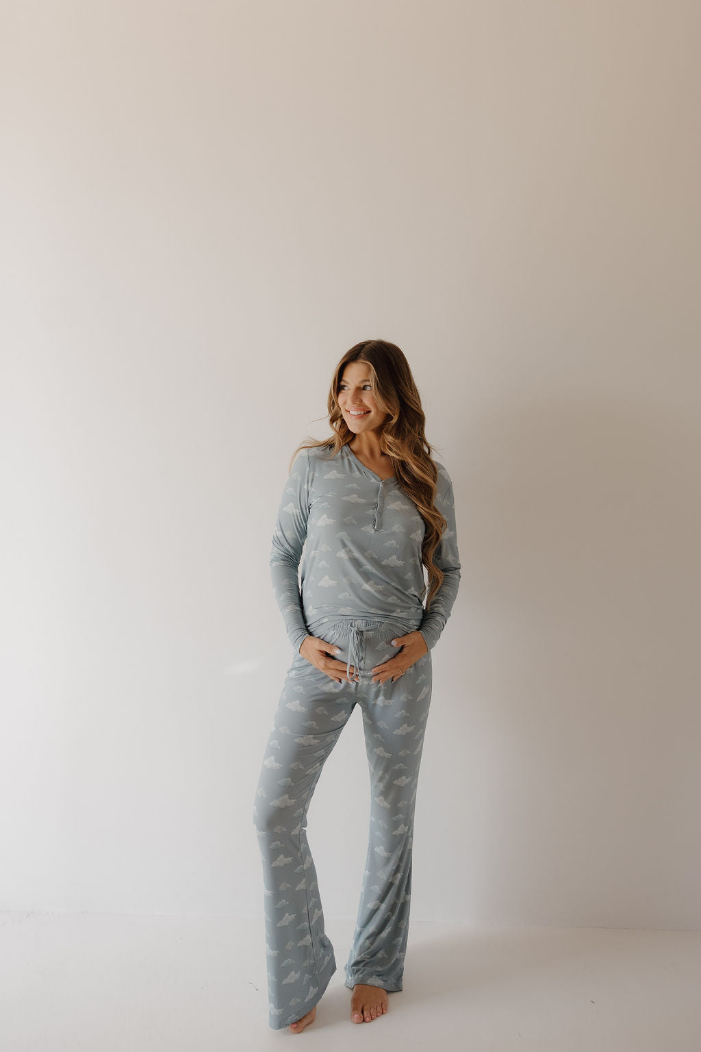 A smiling woman with long hair stands barefoot by a plain light wall, wearing Kendy 3's Women's Bamboo Pajama | Duke in light blue with a subtle white print, gently cradling her belly and enjoying the hypo-allergenic softness.
