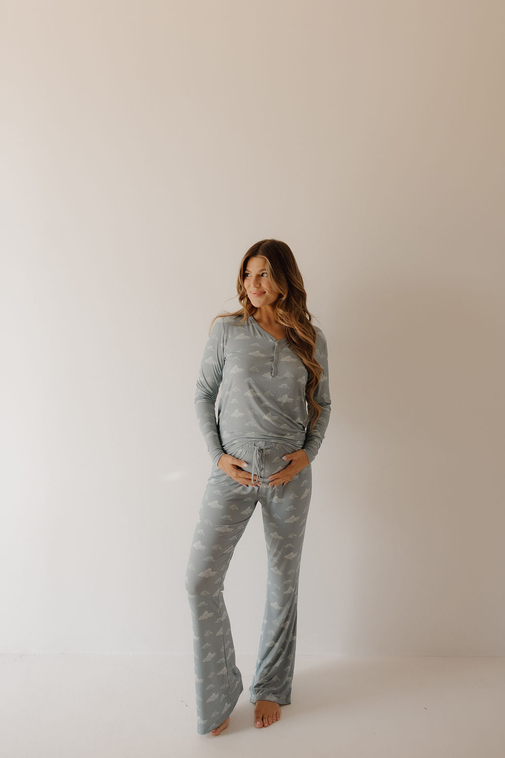 A woman with long hair stands barefoot against a plain background, wearing the Kendy 3 Women's Bamboo Pajama | Duke set in light blue with subtle patterns. One hand on her hip, she gazes to the side, smiling softly.