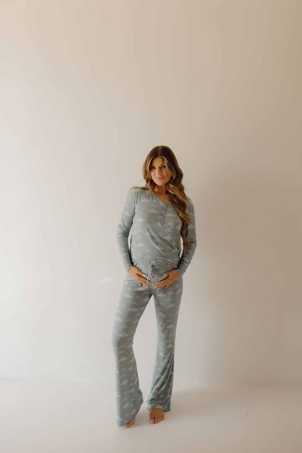 A woman with long wavy hair stands barefoot against a plain light wall, smiling softly with one hand on her hip as she wears the Kendy 3 Women's Bamboo Pajama | Duke set in light blue with a subtle print.