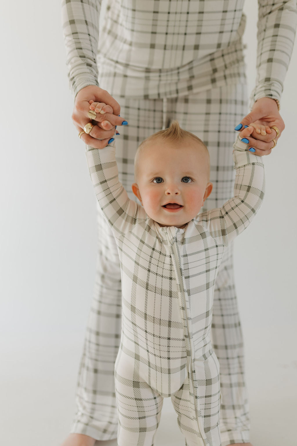 A baby in breathable Bamboo Zip Pajamas by forever french baby, featuring Picnic Plaid design, holds hands with an adult in matching plaid. The smiling baby has a tiny mohawk as they pose against a white background, highlighting the charm of hypoallergenic clothing for little ones.