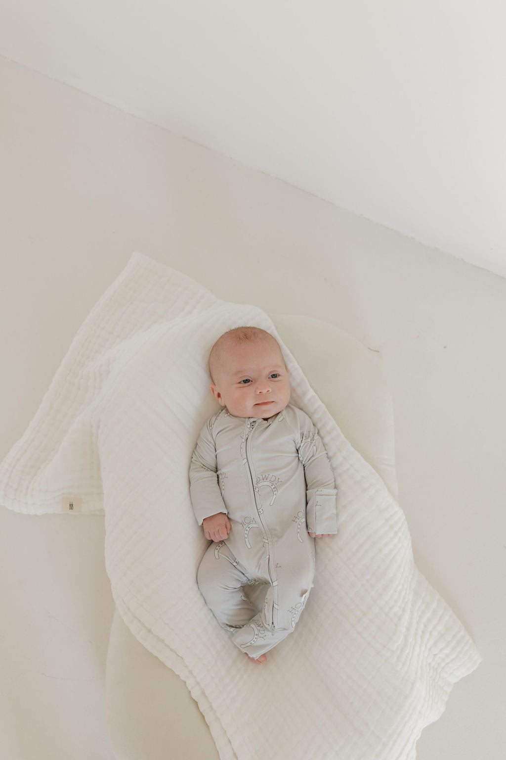 A baby in a light-colored Bamboo Zip Pajamas by forever french baby lies on a white blanket, gazing upward. The minimal white background creates a soft, serene atmosphere enhanced by the hypo-allergenic clothing that ensures delicate comfort.