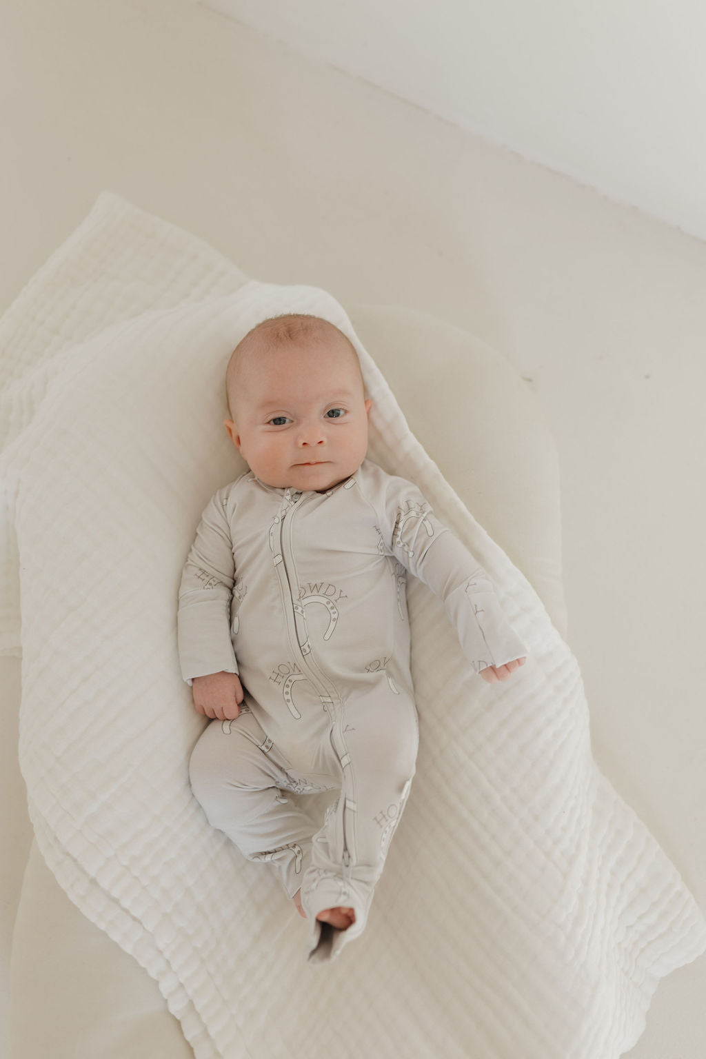 A baby in forever french baby's Bamboo Zip Pajamas | Howdy lies on a white, quilted blanket. The hypo-allergenic outfit ensures comfort as the baby gazes up with a slight smile, enveloped in gentle lighting.