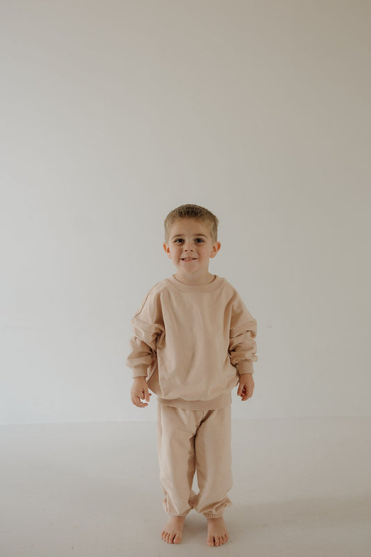 A young child with short light brown hair smiles at the camera, barefoot on a white floor, dressed in the forever french baby Child Sweat Set | Cashew—perfect for cozy, everyday adventures against a clean, light background.