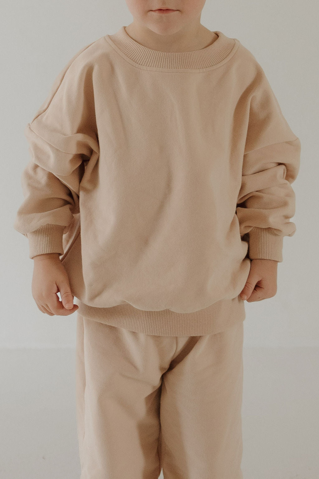 A young child models the forever french baby Child Sweat Set in Cashew against a simple light background, with their face partly out of frame and hands relaxed at their sides.