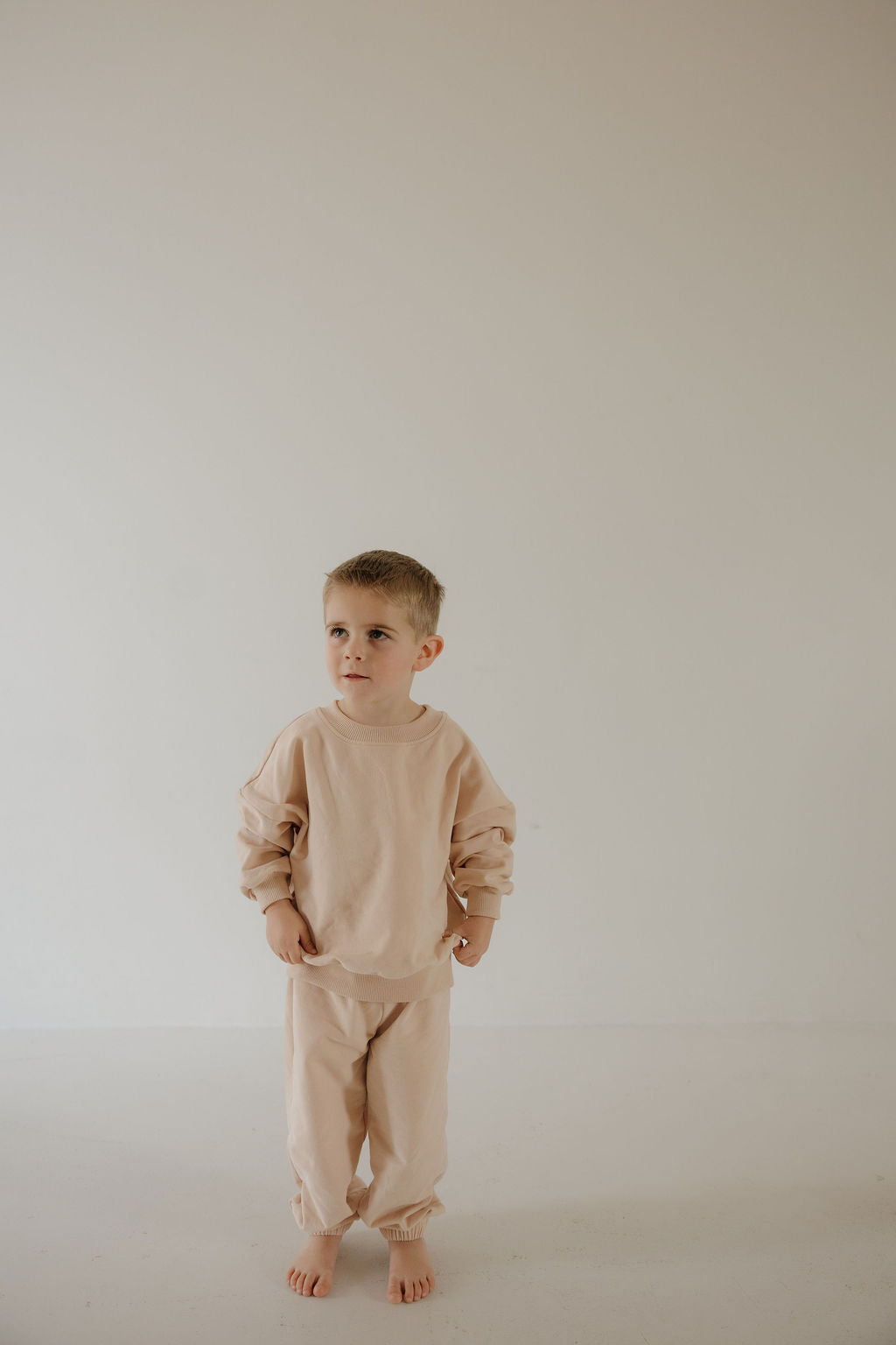 A young boy stands barefoot against a plain wall, wearing the forever french baby Child Sweat Set in Cashew. He gazes to the side with a neutral expression, ready for everyday adventures.