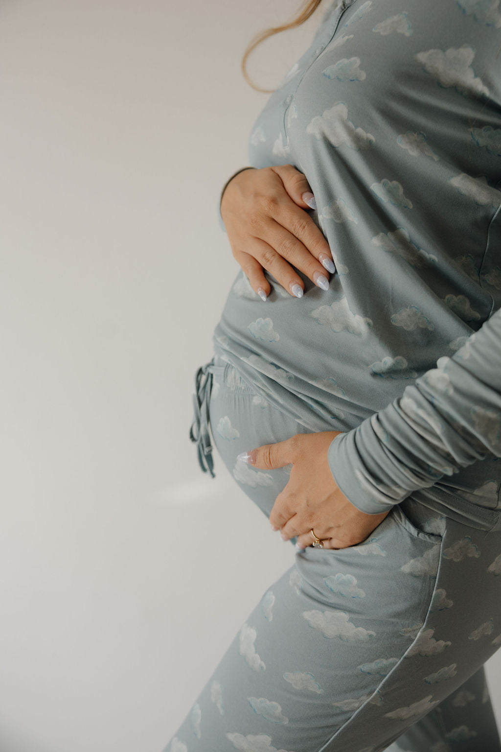 A pregnant person in Kendy 3 Women's Bamboo Pajama | Duke with a cloud pattern holds their belly with both hands, standing against a plain light background.