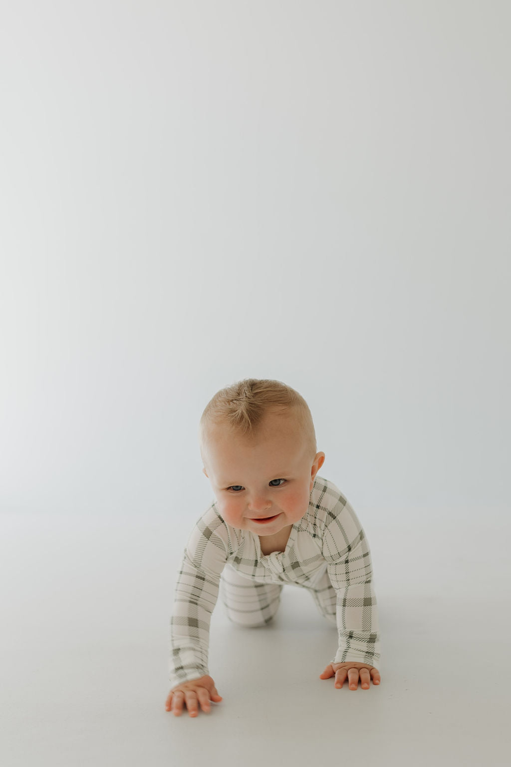 In a plain white setting, a curious baby in "Picnic Plaid" Bamboo Zip Pajamas by forever french baby crawls on a light floor. This breathable onesie ensures comfort as they explore their world.