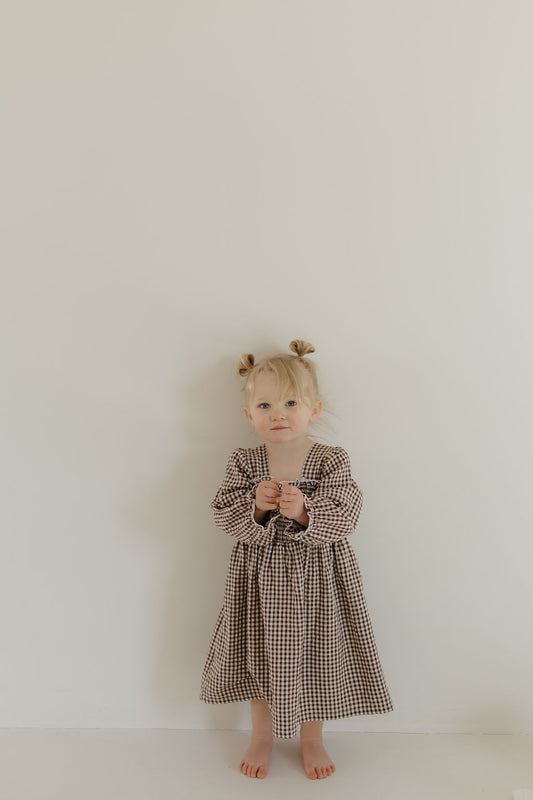 A young child with blonde hair in small pigtails stands against a plain, light-colored wall. They are barefoot and holding something small in their hands while wearing a forever french baby long-sleeved dress in the Chocolate Chai pattern, featuring baby sizes in brown and white checks.