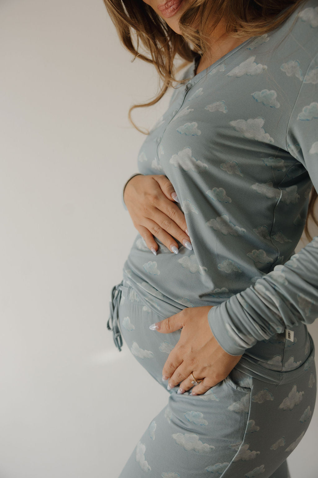A woman, partially out of frame, gently cradles her baby bump while wearing Kendy 3's Women's Bamboo Pajama | Duke in soft blue with a cloud pattern, showcasing its breathable comfort perfect for pregnancy.