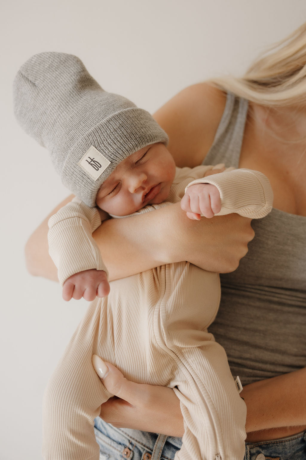 A woman in a gray tank top holds a sleeping baby dressed in forever french baby’s Bamboo Zip Pajamas | Ribbed Cream and a large gray knit hat—a peaceful, relaxed moment.
