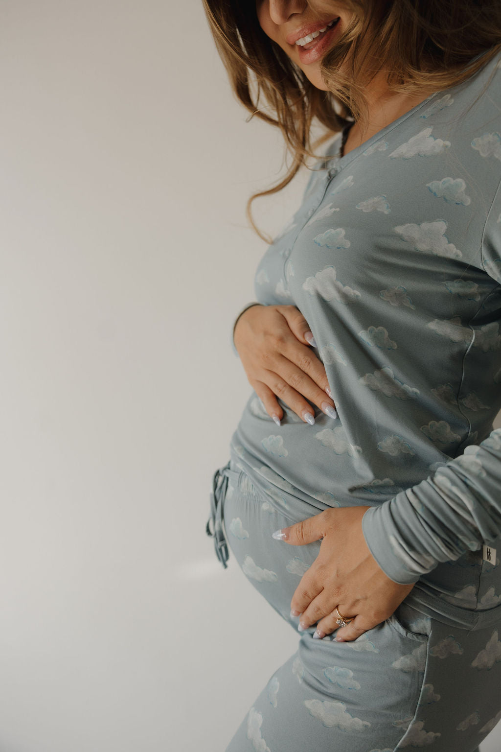 A pregnant woman in Kendy 3's Women's Bamboo Pajama | Duke with a cloud pattern smiles and gently cradles her belly against a plain background.