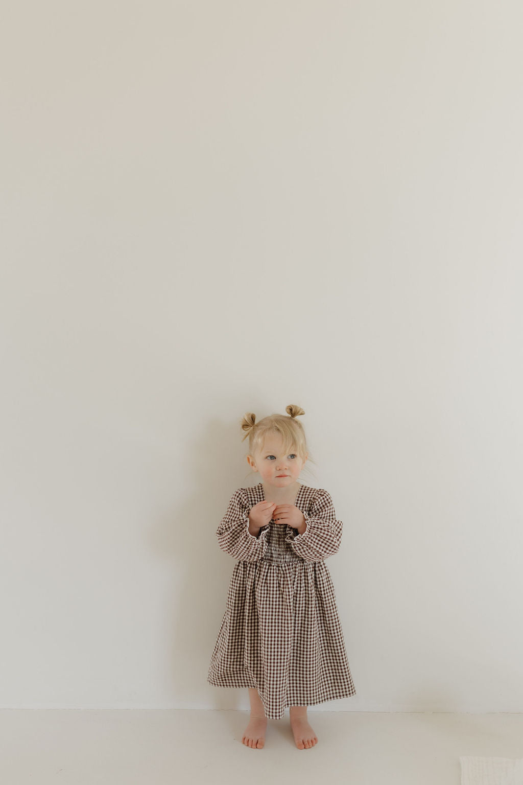 A young child in a forever french baby Child Long Sleeve Dress | Chocolate Chai stands barefoot before a white wall, styling their hair in two small buns while holding the dress front, which pairs with matching rompers for chic sibling coordination.