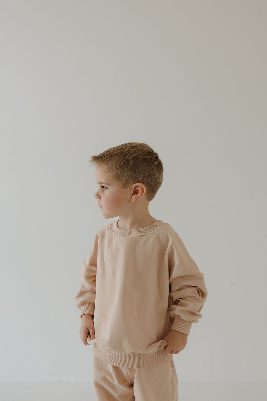 A young boy with light brown hair models the forever french baby Child Sweat Set in Cashew, standing with hands in his pockets against a plain background—ideal for everyday adventures.