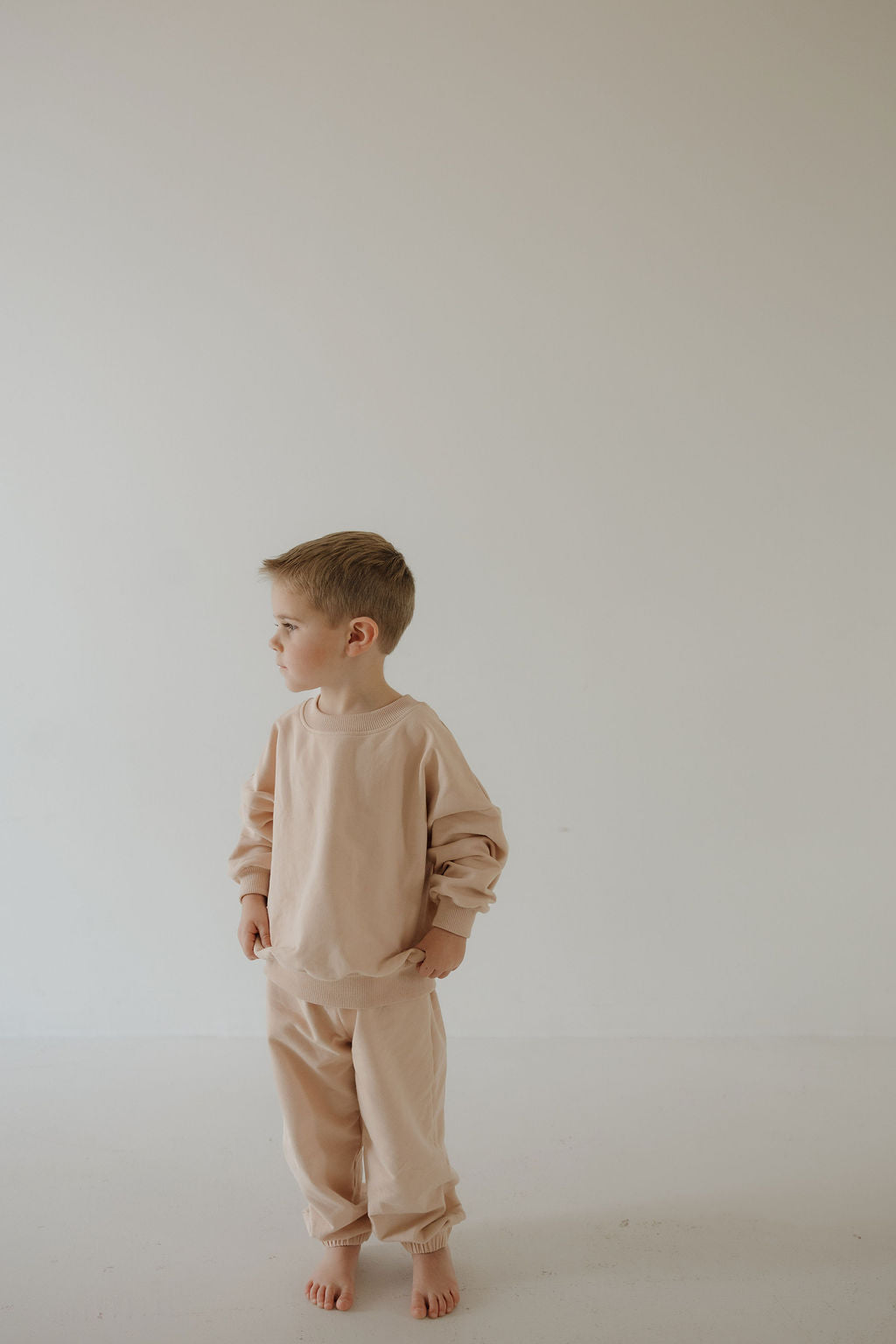 A young child models the forever french baby Child Sweat Set in Cashew, standing barefoot against a light wall with hands in pockets—the simple setting emphasizing everyday charm.