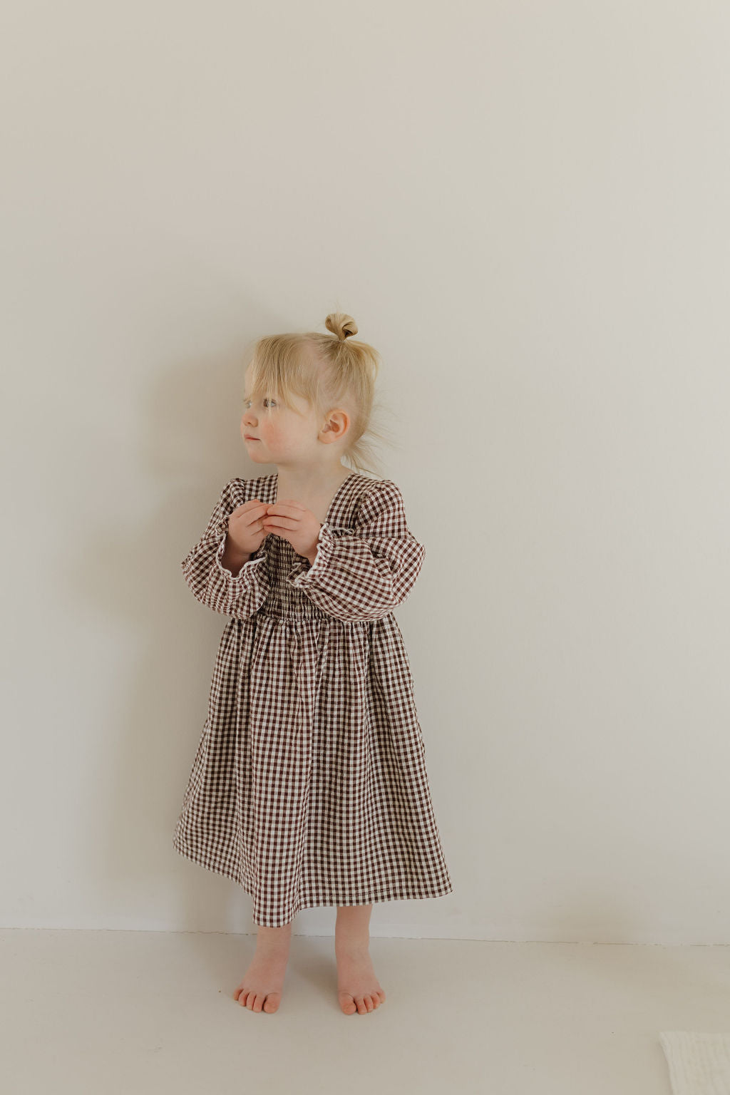 A young child with light hair in a bun stands barefoot against a plain wall, wearing the forever french baby Child Long Sleeve Dress in Chocolate Chai print. They look to the side with hands clasped.