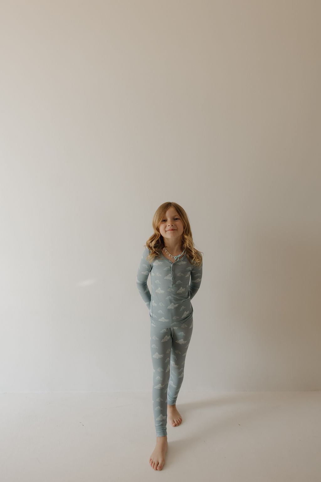 A young girl with wavy blonde hair stands barefoot against a light wall, smiling slightly with her hands behind her back, wearing Kendy 3 Bamboo Two Piece Pajamas | Duke featuring a white cloud pattern.