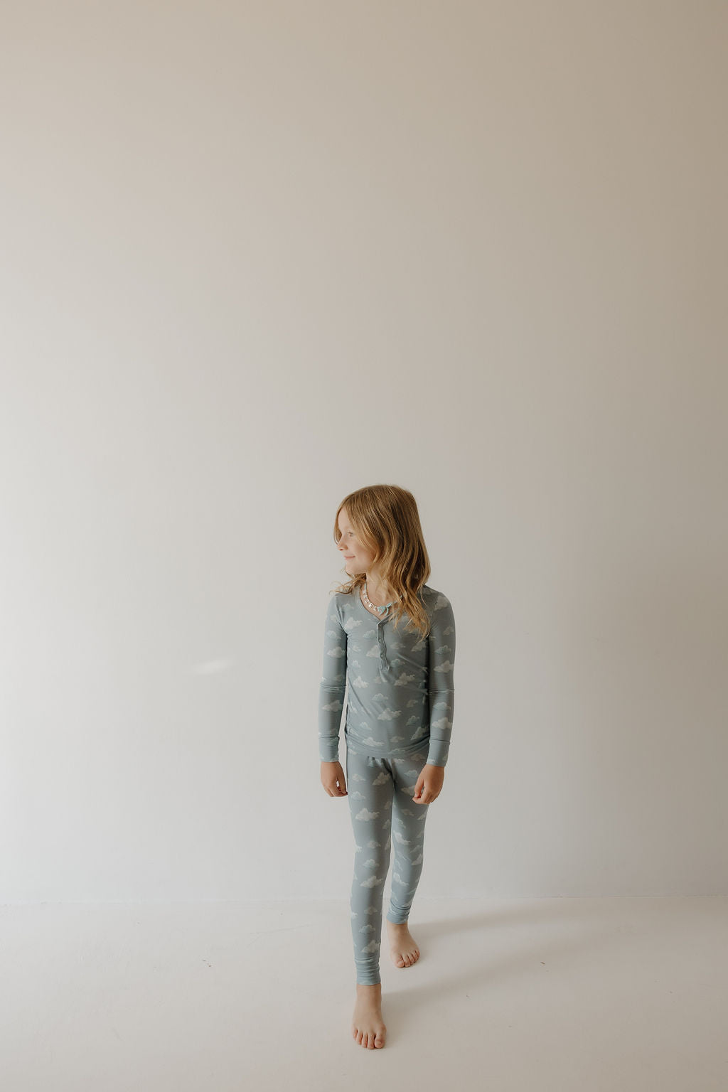 A young blonde girl stands barefoot against a light background, wearing Kendy 3's Bamboo Two Piece Pajamas | Duke in a matching light blue set with a subtle white pattern, looking left with a relaxed expression.