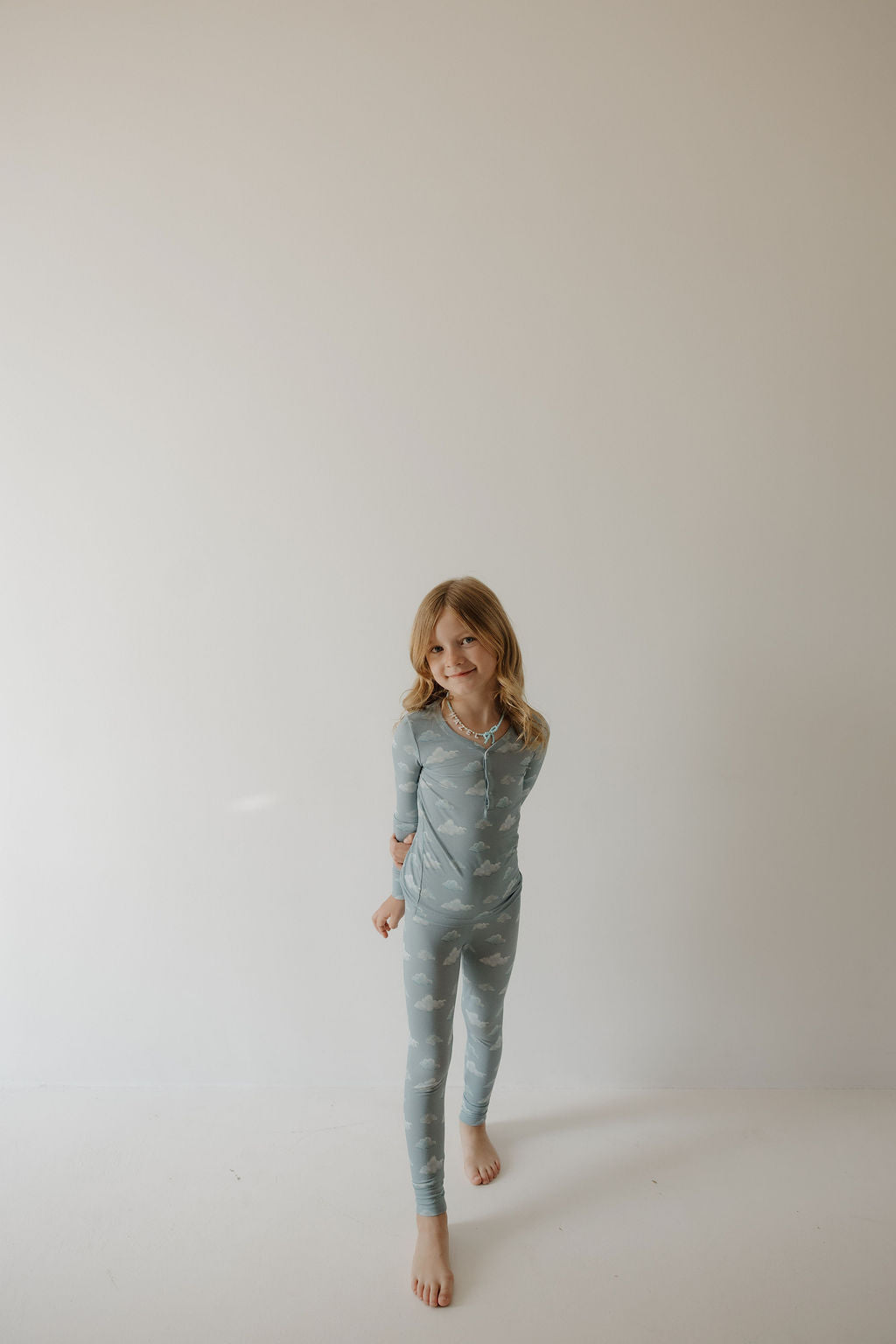 A young child with long blonde hair stands barefoot against a plain light background, wearing Kendy 3 Bamboo Two Piece Pajamas | Duke in a light blue cloud pattern and smiling slightly at the camera.