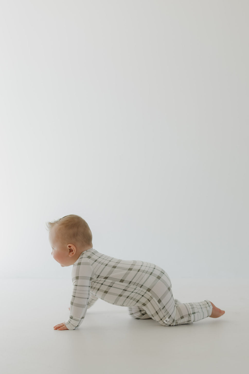 A baby in a Bamboo Zip Pajamas from forever french baby, with Picnic Plaid design and long sleeves, is shown in profile, crawling on all fours against a plain light background. The baby has short, light-colored hair.