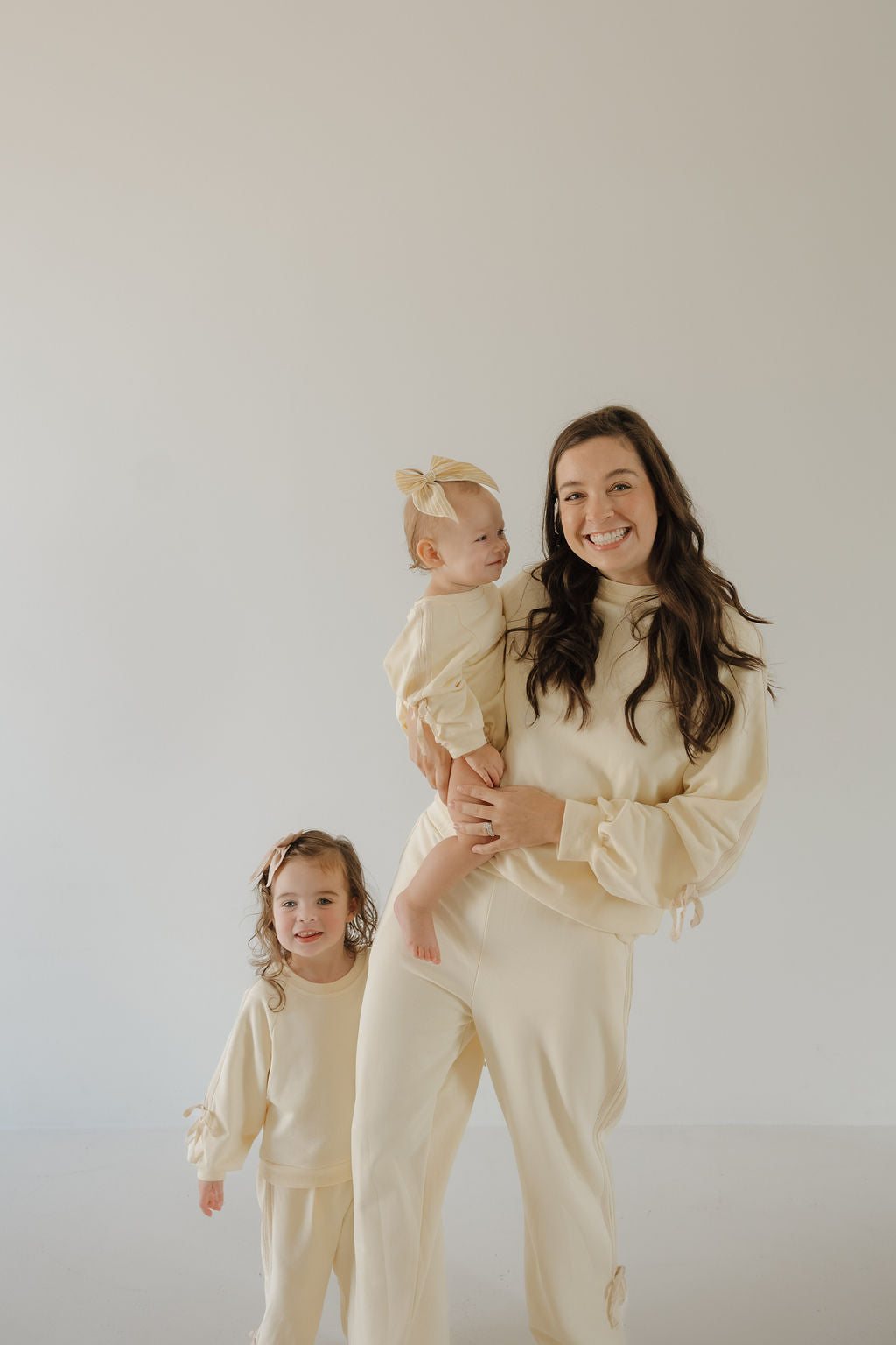 A woman in the forever french baby Women's Sweat Set | Buttercream Bows smiles while holding a baby in a cozy romper next to a young girl; all wear matching light beige outfits against a simple, light background.
