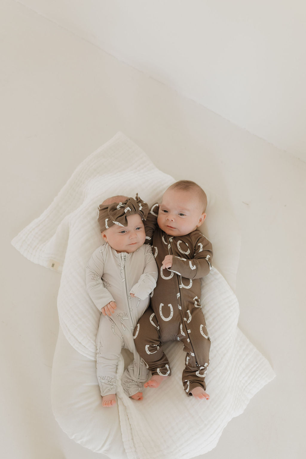 Two babies lie on a soft white blanket. The baby on the left wears a light-colored onesie with leaf patterns and matching headband, made from hypo-allergenic fabric. The baby on the right wears a dark "Bamboo Zip Pajamas | Howdy" onesie with white crescents by forever french baby. Both gaze up.