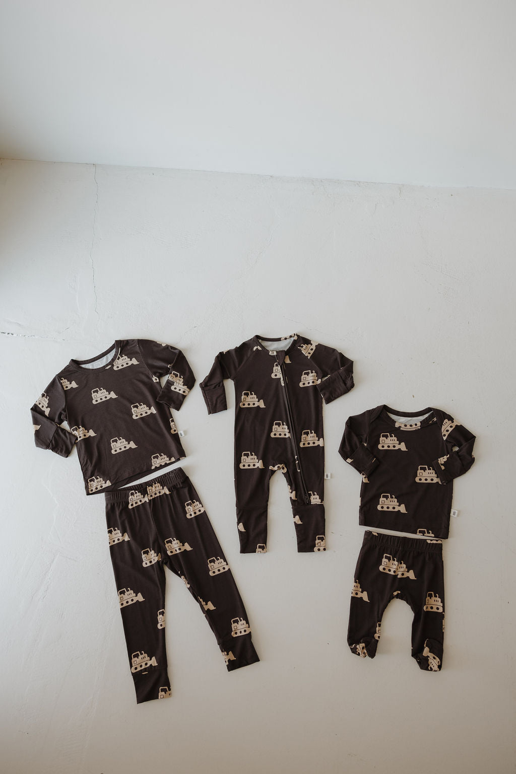 Four matching Sleepy Time Set | Bulldozer pajamas by forever french baby, in dark brown bamboo with tan castle print, are laid out on a light floor. Includes two long-sleeve tops with pants and two long-sleeve onesies with pants.