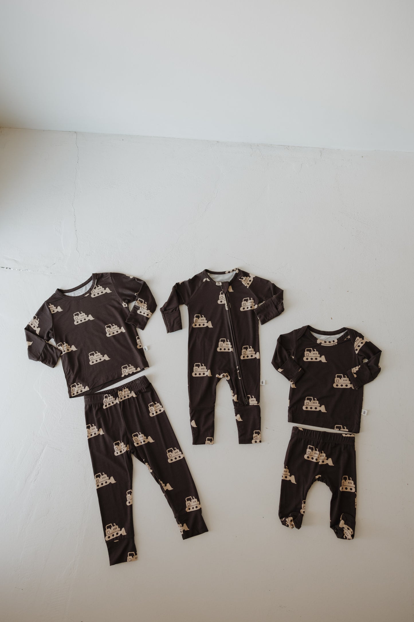 Four matching dark brown Sleepy Time Set | Bulldozer outfits from forever french baby, featuring a beige truck pattern, are arranged on a white floor. The set includes two pajama sets and two onesies in breathable, hypoallergenic fabric.