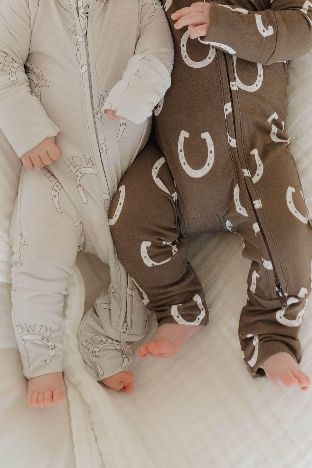 Two babies rest on a soft, white blanket. One wears a light beige onesie with dark horseshoes, the other dons a dark brown one with light horseshoes. Their forever french baby Bamboo Zip Pajamas | Howdy are ideal for delicate skin, keeping them snug and comfy all day long.