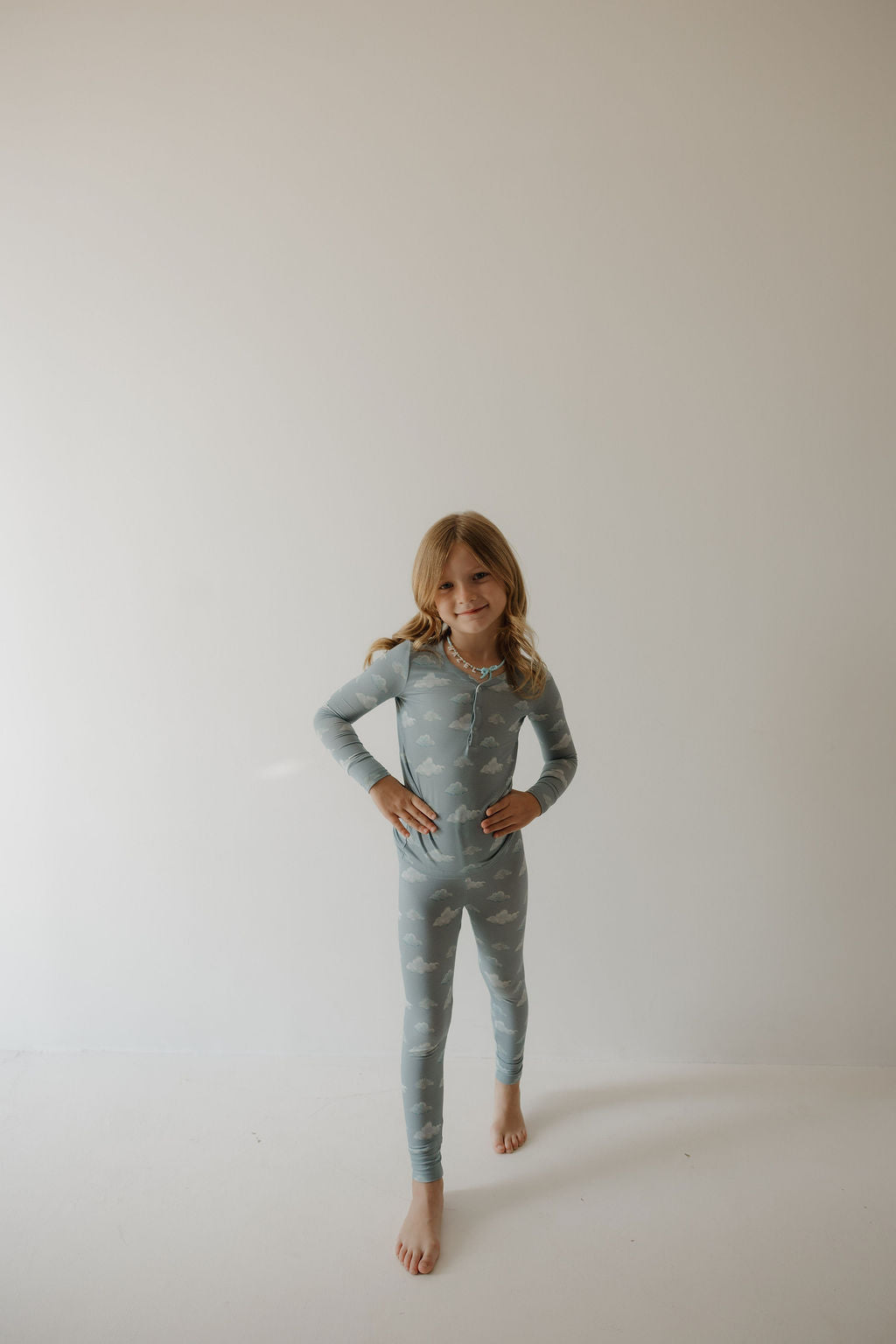 A young girl stands barefoot on a white floor, smiling with hands on hips, wearing the Kendy 3 Bamboo Two Piece Pajamas | Duke in light blue with a cloud pattern against a plain light background.