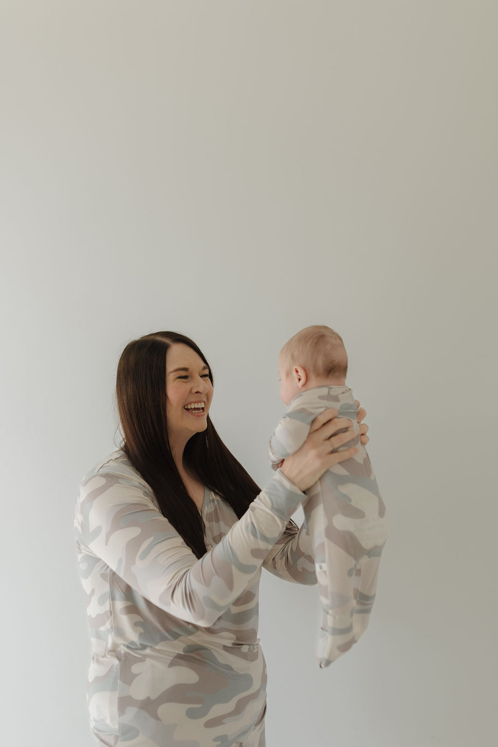 A woman smiles as she holds her baby aloft, both wearing matching Bamboo Zip Pajamas in Camo by forever french baby against a light backdrop. The hypoallergenic design enhances their comfort, adding to the picture-perfect moment.