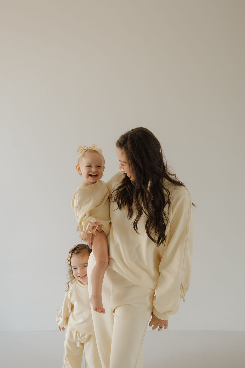 A woman in the forever french baby Women's Sweat Set | Buttercream Bows smiles at a little girl on her hip, with another young girl beside them; all three wear matching sets in a light, minimalist space.