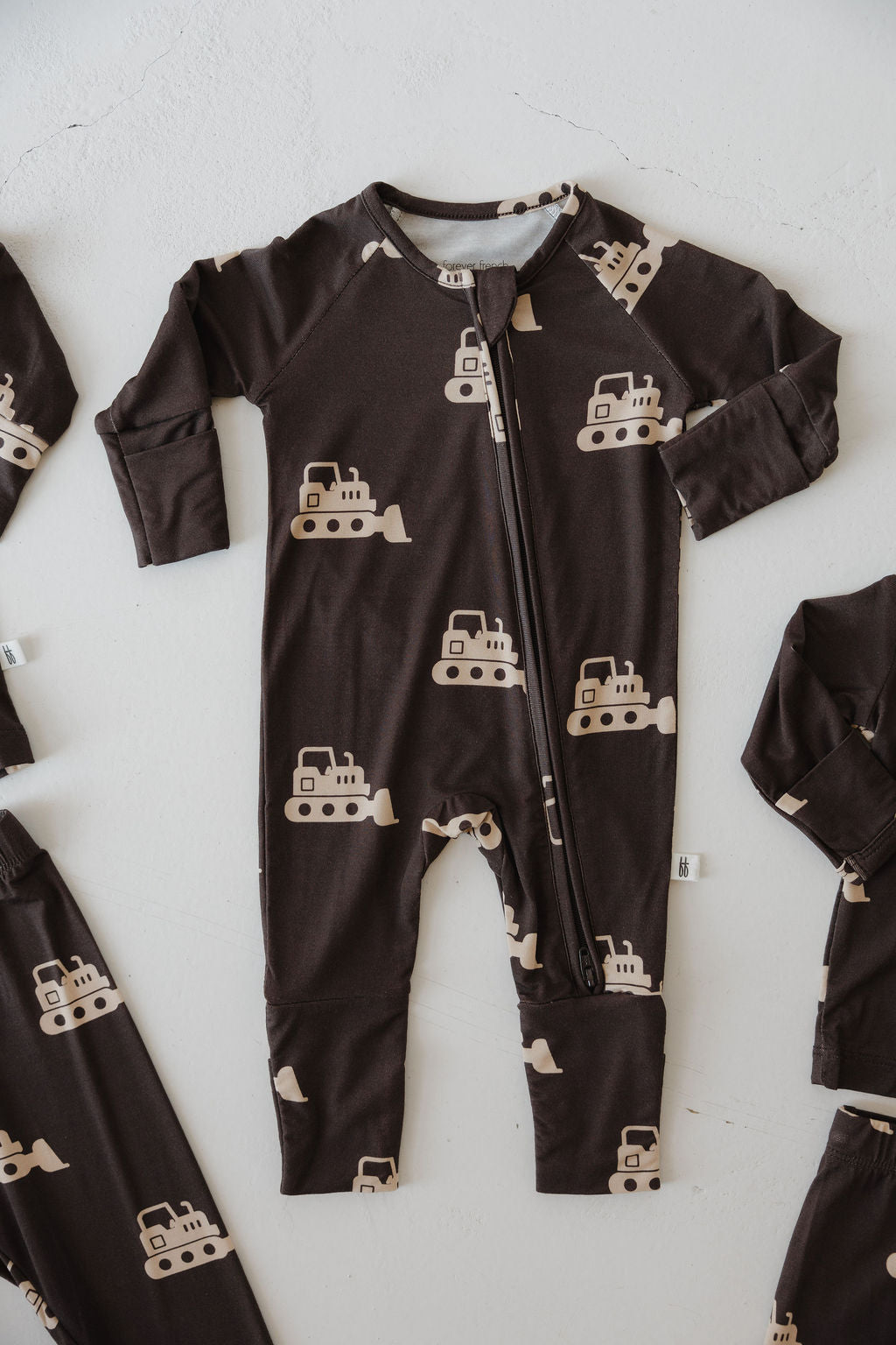 The forever french baby Bamboo Zip Pajamas | Bulldozer is a dark brown, hypo-allergenic onesie featuring beige construction bulldozers and a zipper, shown laid flat on a white surface.