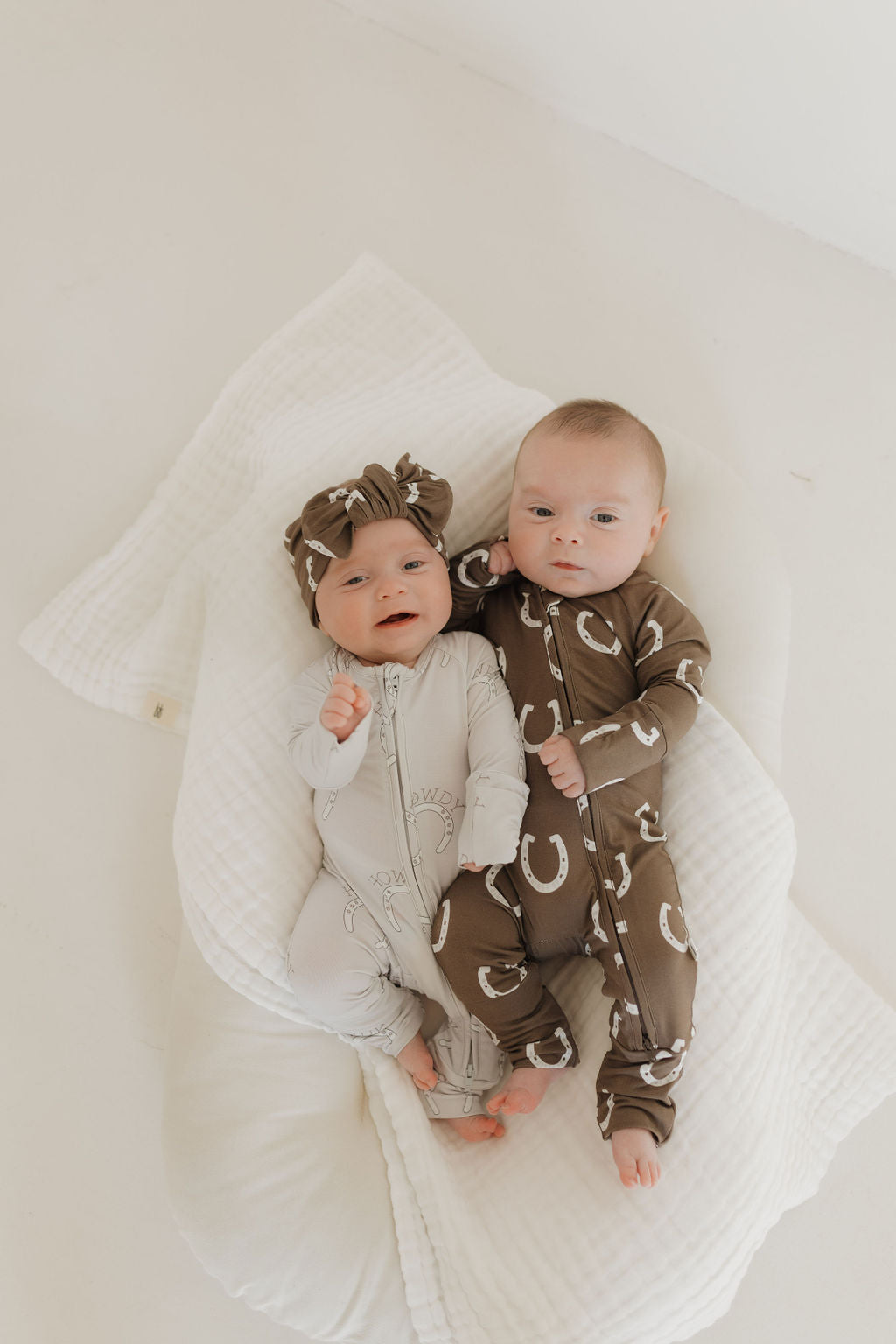 Two infants are lying on a soft, white cushion. One is wearing light-colored onesie and matching headband, while the other wears a hypo-allergenic brown Bamboo Zip Pajamas from forever french baby with white patterns. Both have relaxed expressions as they look up.