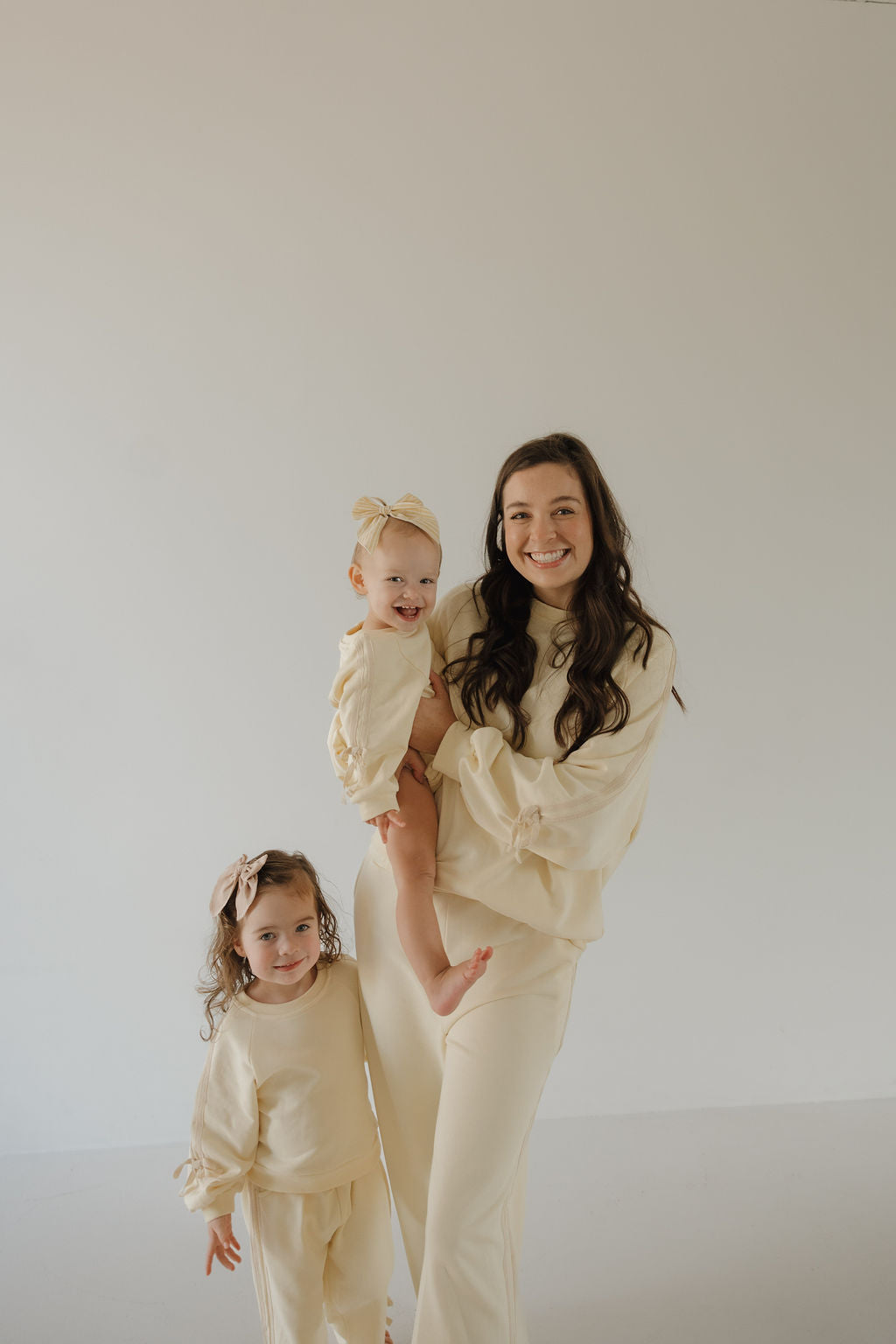 A woman in the Women's Sweat Set | Buttercream Bows by forever french baby smiles while holding a baby in a matching romper, next to a young girl; all three wear coordinated cream outfits and headbands against a simple backdrop.